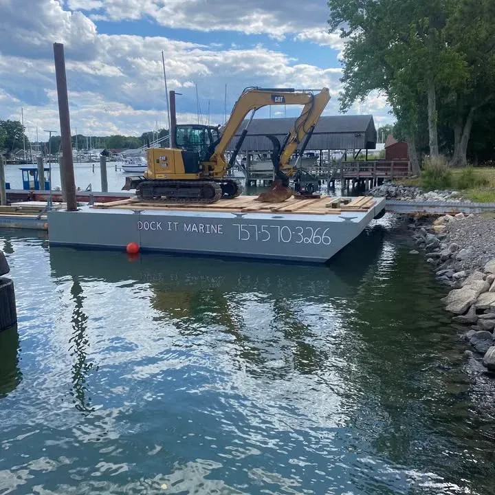 The Dock It Marine work barge, with a CAT excavator on deck and DOCK IT MARINE 757-570-3266 painted on the hull, beside a rip-rap shoreline and marina